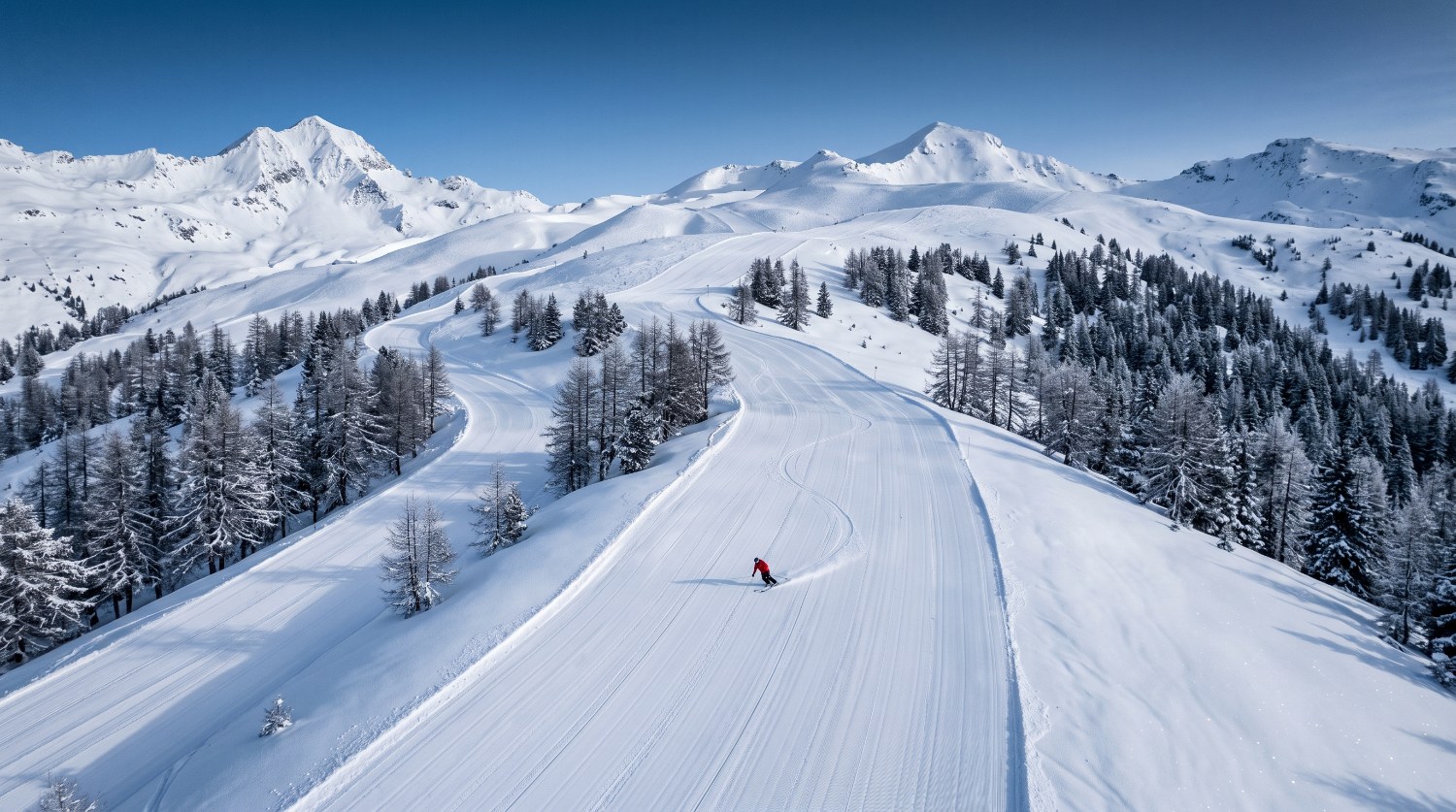 Ampie piste appena battute tra larici innevati, una persona in rosso traccia le prime curve